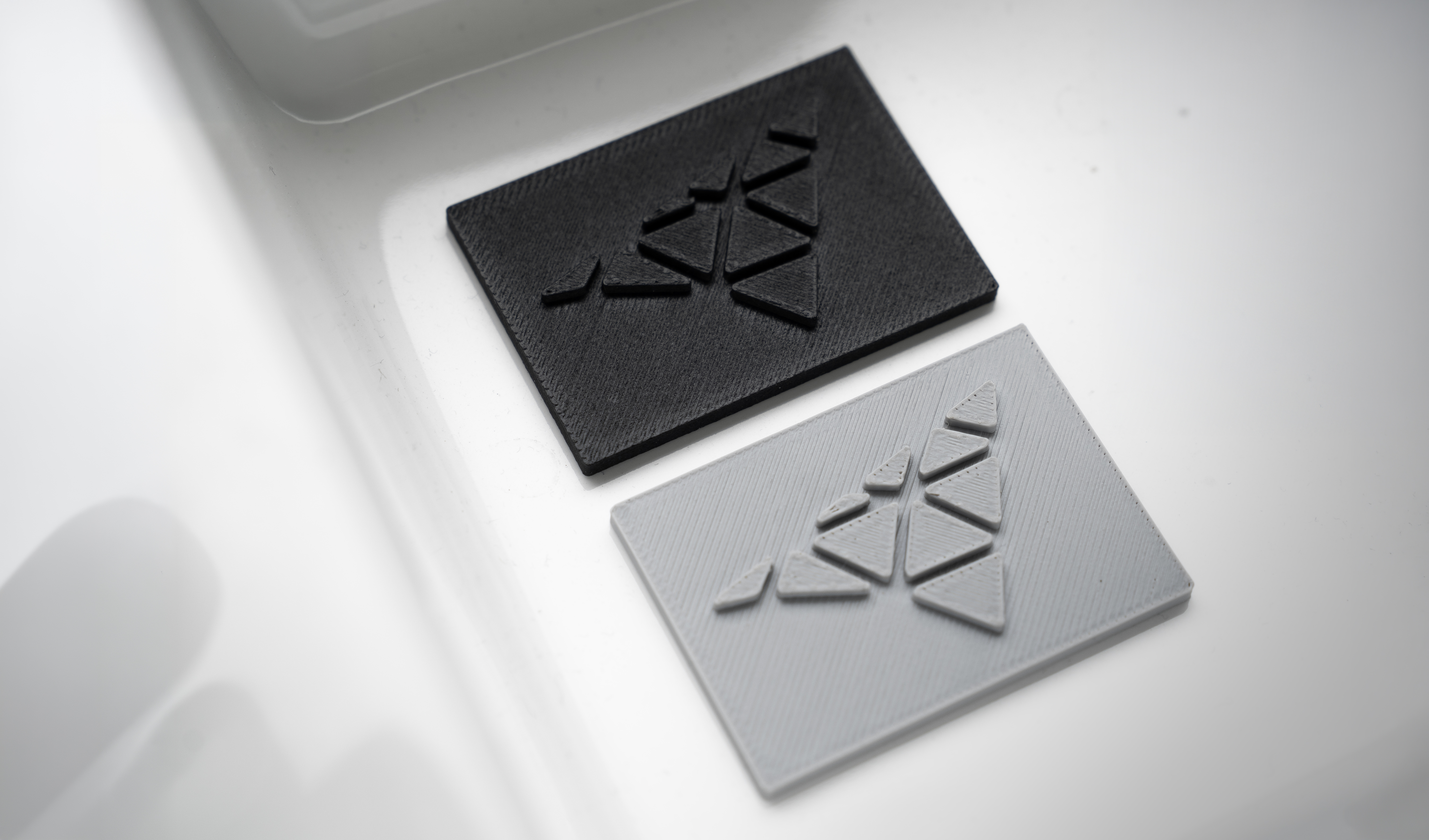 Zwei FDM-gedruckte Musterplättchen in Schwarz und Grau mit Logo